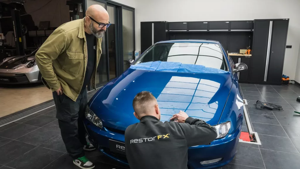 Gerry de l’émission Wheeler Dealers sur RMC Découverte observe Mehdi, technicien RestorFX, préparant la Peugeot 406 Coupé V6 avant l’application de la technologie de rénovation sans peinture RestorFX.